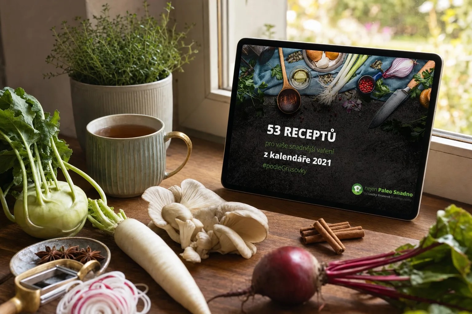 E-kniha 53 receptů z kalendáře 2021 na tabletu na dřevěném parapetu u okna, vedle hrnek čaje, kedlubna, hlíva ústřičná, bílá ředkev, červená řepa, badyán a skořice