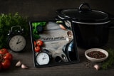 Kuchařka Vaříme pomalu na tabletu opřeném o černý pomalý hrnec, vedle budík se dvěma zvonky, tmavé studio
