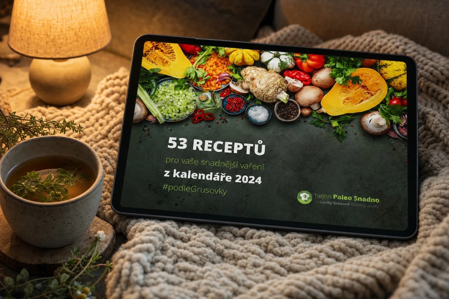 E-kniha 53 receptů z kalendáře 2024 na tabletu položeném na pletené dece na pohovce ve večerním světle stolní lampy, vedle hrnek bylinkového čaje na dřevěném podtácku