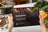 Ruce drží tablet s e-knihou 53 receptů z kalendáře 2022 nad kuchyňskou linkou, vzadu hrnec s bublající polévkou, kolem zázvor, chilli papričky, česnek, kadeřávek a půlka dýně hokkaido