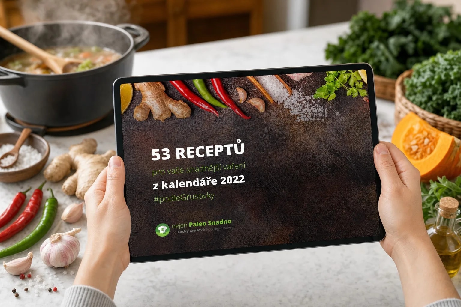 Ruce drží tablet s e-knihou 53 receptů z kalendáře 2022 nad kuchyňskou linkou, vzadu hrnec s bublající polévkou, kolem zázvor, chilli papričky, česnek, kadeřávek a půlka dýně hokkaido